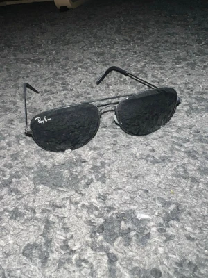 Svarta Ray-Ban aviator solglasögon - Säljer ett par klassiska svarta aviator solglasögon från Ray-Ban. De har tunna metallbågar och mörka linser som ger en tidlös och cool look. Perfekta för att lyfta vilken outfit som helst och skydda ögonen mot solen.