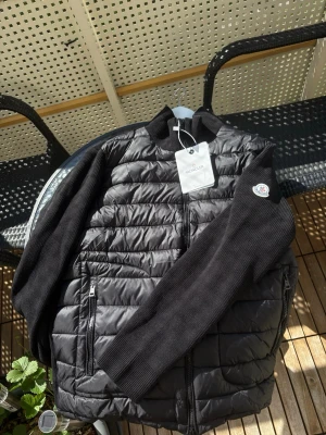 Svart cardigan från Moncler - Snygg svart cardigan från Moncler med quiltad front och stickade ärmar. Jackan har dragkedja framtill, två sidofickor med dragkedja och klassisk Moncler-logga på ärmen. Perfekt för kalla vinterdagar och riktigt stilren look. Storleken på lappen är XS men passar inte ens i närheten, är mer som en L. Aldrig använd med alla lappar kvar