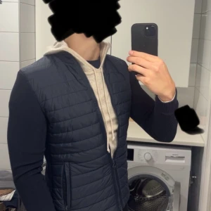 Mörkblå cardigan jacka från J.Lindeberg  - Snygg mörkblå quiltad jacka från J.Lindeberg med ribbad krage och ärmar. Jackan har dragkedja framtill, två sidofickor med dragkedja och diskret logga på ena ärmen. Materialmix med stickade ärmar och quiltad front ger en modern look. Storlek S. Hör av dig vid frågor!