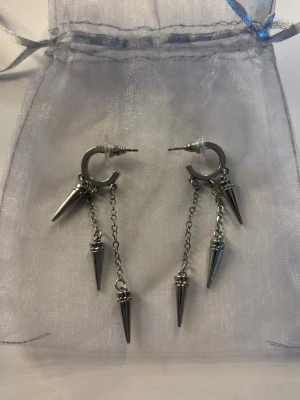 Silvriga örhängen med kedjor och spikes - Säljer ett par coola silvriga örhängen med ringar och hängande kedjor med spetsiga spikes. Örhängena har en edgy vibe och är tillverkade i metall med flera detaljerade hängen. Perfekta för dig som vill sticka ut och addera lite attityd till din stil. Helt oanvända men har tyvärr inte kvar förpackningen de satt på 💕