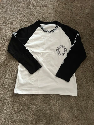 Svartvit långärmad topp Chrome Hearts - Säljer en svartvit långärmad topp från Chrome Hearts med tryck både fram och bak samt logga längs halsringningen och ärmarna. Klassisk basebollmodell med kontrastfärgade ärmar och rund hals. Perfekt för dig som gillar streetwear och vill sticka ut.