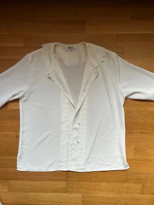 Vit blus med scallop-krage från RIO - Stilren vit blus från RIO med långärmad design och unik scallop-detaljerad krage. Framsidan har dekorativa veck och knäppning. Perfekt för dig som gillar klassiska men ändå lekfulla detaljer. Materialet känns mjukt och har ett lätt glansigt utseende.