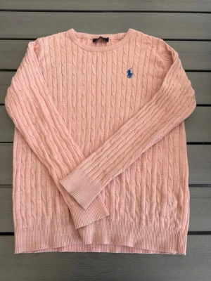 Ljusrosa kabelstickad tröja Polo Ralph Lauren - Supersnygg ljusrosa kabelstickad tröja från Polo Ralph Lauren med blå logga broderad på bröstet. Tröjan har rund halsringning, långa ärmar och ribbade muddar. Perfekt för dig som gillar klassisk och stilren look. knappast använd, priset går o diskutera vid snabb affär! 