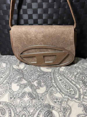 Beige handväska  - Snygg beige handväska  i tygmaterial med ett stort metallfärgat D-logotyp på framsidan. Väskan har axelrem och stängs med lock. Perfekt för dig som gillar streetstyle och vill ha något unikt.