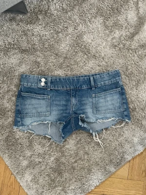 Blå jeansshorts med råa kanter - Säljer ett par blå jeansshorts med slitna, råa kanter och stora framfickor. Shortsen har dubbla knappar i midjan, bälteshällor och bakfickor. Perfekta för en avslappnad och trendig sommarlook. Saknas en knapp men inget som stör funktionen (den var borta när jag köpte dom)