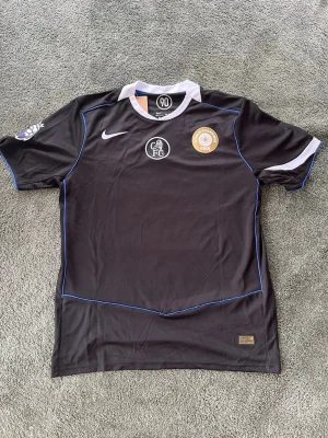 Svart Nike fotbollströja Chelsea - Snygg svart fotbollströja från Nike med Chelsea FC-logga och FIFA World Champions-märke på bröstet. Tröjan har vita detaljer vid kragen och ärmsluten samt tunna blå linjer längs sidorna. Tillverkad i lätt och ventilerande material, perfekt för träning eller match.