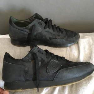 Svarta sneakers från Philippe Model - Pris kan diskuteras. Snygga svarta sneakers från Philippe Model i storlek 43. Skorna har diskret kamouflagemönster, svarta skosnören och en klassisk gummisula. Tillverkade i skinn och textil med coola detaljer och logga på sidan. Perfekta för dig som gillar stilrena och unika sneakers.