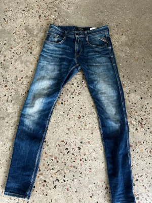 Blå Replay jeans med slitningar - Säljer ett par blå jeans från Replay med snygga slitningar och tvättade detaljer. Klassisk femficksmodell med smal passform och kontrastsömmar. Materialet är denim i bomull och byxorna har en modern, trendig look.