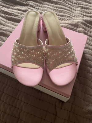 Rosa sandaler med pärldetaljer - Säljer ett par ljusrosa sandaler med öppen tå och låg klack. Ovandelen är dekorerad med pärlor och små glittriga stenar på ett nätliknande material. Snygg och feminin design som sticker ut. Perfekta för dig som vill ha något extra till din outfit.
