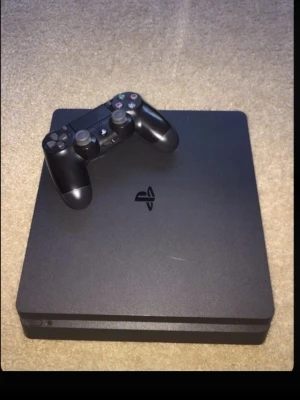 PlayStation 4 Slim med handkontroll - Säljer en svart PlayStation 4 Slim med tillhörande svart DualShock 4-handkontroll. Konsolen har en stilren, fyrkantig design och är tillverkad i plast. Perfekt för gaming och streaming.