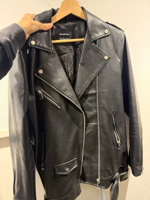 Svart skinnjacka - Snygg svart skinnjacka med klassisk bikerstil. Jackan har silvriga dragkedjor, snedställd front, axelklaffar och bälte i midjan. Perfekt för dig som vill ha en tidlös och cool look.