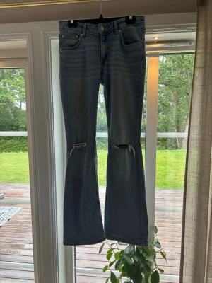 Blå bootcut jeans med slitna knän - Säljer ett par blå bootcut jeans med slitningar över knäna och klassisk femficksdesign. Jeansen har låg midja och är tillverkade i denim med lätt stretch. Perfekta för dig som gillar en avslappnad men trendig look.