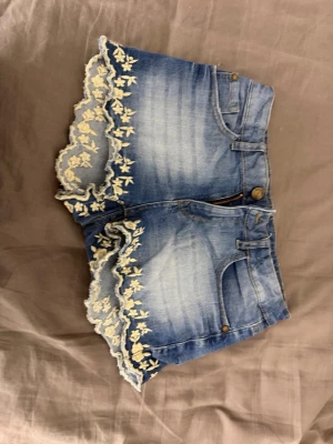 Blå jeansshorts med spetsdetaljer - Snygga blå jeansshorts med ljus tvätt och dekorativa vita spetsdetaljer längs bensluten. Shortsen har klassisk femficksdesign, knapp och dragkedja framtill samt en feminin touch med blommig spets. Perfekta för sommardagar!