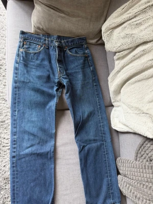 Blå raka jeans från Levi's - Klassiska blå jeans från Levi's med rak passform och fem fickor. Jeansen har en tidlös look med mellanblå tvätt och detaljer som kopparfärgade nitar och läderpatch bak i midjan. Perfekta för en avslappnad och stilren outfit.