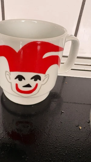 Vit mugg med röd clownmotiv - Unik vit mugg i keramik med ett stort rött clownansikte tryckt på framsidan. Muggen har ett rejält handtag och rymmer mycket dryck. Perfekt för dig som gillar roliga och färgglada detaljer i köket.