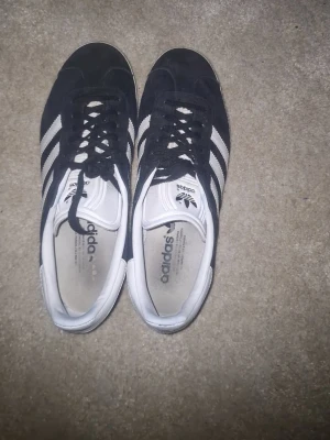 Svarta Adidas sneakers med vita ränder - Klassiska svarta Adidas sneakers med tre vita ränder på sidorna och vit hälkappa med Adidas-logga. Skorna har svart mockaliknande material och vita detaljer på plös och insida. Lågt skaft och snörning framtill ger en tidlös streetstil.