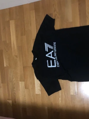 Svart EA7 Emporio Armani t-shirt - Snygg svart t-shirt från EA7 Emporio Armani med stor vit logga framtill. Klassisk passform och rund halsringning. Perfekt för dig som gillar stilrena och trendiga plagg med tydlig branding.