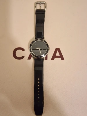 Svart klocka från Marc Jacobs - Stilren svart klocka från Marc Jacobs med rund urtavla och silverdetaljer. Klockan har ett svart läderband och silverfärgat spänne. Urtavlan är enkel med diskreta markeringar och logga. Perfekt accessoar för en clean look.
