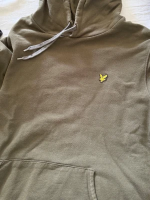 Olivgrön hoodie från Lyle & Scott - Olivgrön hoodie från Lyle & Scott med klassisk gul logga på bröstet. Hoodien har huva med dragsko och en stor magficka framtill. Tillverkad i mjukt bomullsmaterial som är skönt att bära. Perfekt för en avslappnad och stilren look.