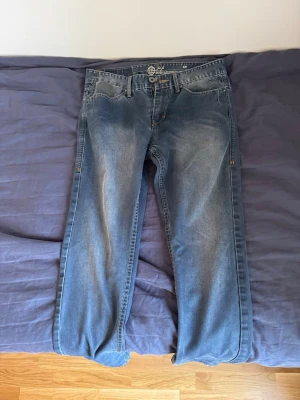 Blå raka jeans med slitningar. W31 - Säljer ett par blå jeans med raka ben och snygga slitningar framtill. Jeansen har klassisk femficksdesign och knappgylf. Materialet är denim i bomull och de har en avslappnad passform som passar till det mesta. Storlek W31