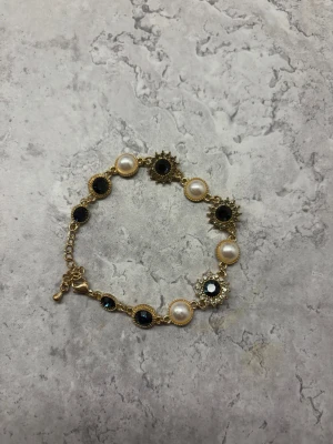 Vintage guldarmband med pärlor och blåa stenar - Snyggt armband i guldfärgad metall med växelvis vita pärlor och blåa glittrande stenar. Armbandet har dekorativa detaljer runt stenarna och justerbar kedja för perfekt passform. Perfekt accessoar för att lyfta din outfit.
