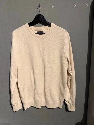Beige stickad tröja  - En stilren beige stickad tröja med rund halsringning och ribbade muddar vid ärmslut och nederkant. Tröjan har en enkel, klassisk design och är perfekt för lager på lager. Passar både killar och tjejer och är lätt att matcha med olika outfits.