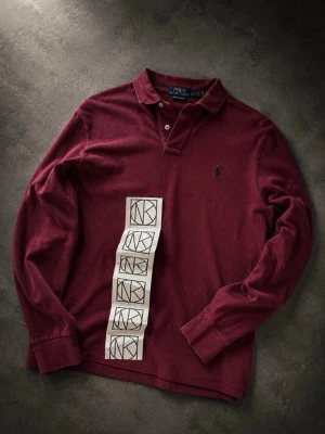Vinröd långärmad piké Polo Ralph Lauren - Vinröd långärmad piké från Polo Ralph Lauren i mjuk bomull. Klassisk krage med två knappar och broderad blå logga på bröstet. Perfekt för dig som gillar stilrena och bekväma plagg. Köpt på Nk kvitto följer med och man får se det på sista bilden. Passar nog s också
