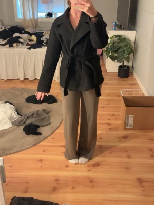 Svart kappa med knytskärp - Stilren svart kappa med bred krage och knytskärp i midjan. Kappan har en klassisk, rak passform och är tillverkad i ett tjockare tyg som passar perfekt för kyligare dagar. Enkel design utan synliga knappar framtill ger ett modernt intryck.