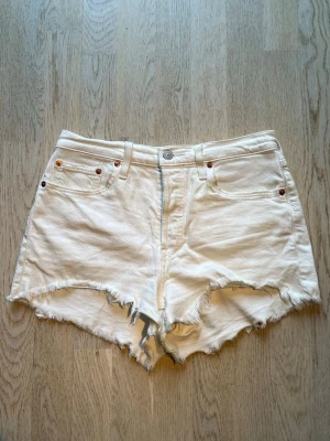 Vita högmidjade jeansshorts Levi's - Säljer ett par vita högmidjade jeansshorts från Levi's med råa, fransiga kanter och klassisk femficksdesign. Shortsen har knappar framtill samt Levi's ikoniska läderpatch bak. Perfekta för sommaren! Tveka inte på att skriva en fråga eller be om fler bilder.🌸🌸