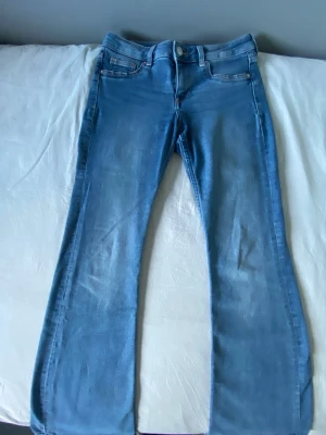 Blå bootcut jeans med hög midja - Säljer ett par blå bootcut jeans med låg midja och klassiska fem fickor. Jeansen har en snygg tvätt och utsvängda ben, perfekta för en trendig och avslappnad stil. Materialet är mjukt denim som sitter bekvämt hela dagen.