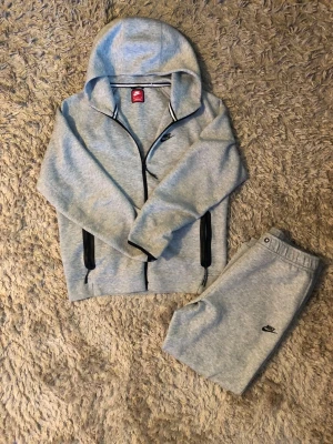 Grå Nike Tech Tracksuit från Nike - Säljer ett par ljusgrå mjukisbyxor från Nike med svart logga på vänster ben och en svart dragkedja på ena fickan. Byxorna har resår i midjan och muddar vid bensluten. Perfekta för chill dagar eller träning.