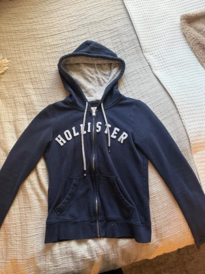 Mörkblå Hollister hoodie med dragkedja - Snygg mörkblå hoodie från Hollister med vit text på bröstet och dragkedja framtill. Har två fickor, vit dragsko i huvan och grått innerfoder i huvan. Perfekt för en avslappnad och trendig look.