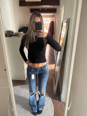 Blå slitna bootcut jeans - Säljer ett par blå bootcut jeans med slitningar över knäna och råa kanter. Jeansen har låg midja och klassisk femficksdesign. Perfekta för en avslappnad och trendig look.