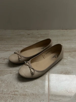 Beige ballerinaskor med rosett - Säljer ett par stilrena beige ballerinaskor med söt rosettdetalj framtill. Skorna har rund tå och är tillverkade i ett slätt material med mjuk innersula för extra komfort. Perfekta för en enkel och clean look.