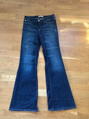 Mörkblå bootcut jeans Gina Jeans - Snygga mörkblå bootcut jeans från Gina Jeans med klassisk femficksmodell och markerade sömmar. Jeansen har utsvängda ben och subtila slitningar framtill för en cool vintagekänsla. Perfekta till sneakers eller boots.