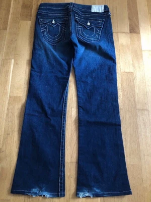 True religion mörkblå bootcut jeans med kontrastsömmar - Snygga true religion mörkblå bootcut jeans med vita kontrastsömmar och detaljerade bakfickor med lock och silverfärgade utstående knappar. Silvrig etikett. Slitna i nederkant (se bild) för övrigt fint skick.  Mått: W30 Innerbenslängd 78 cm Ytterbenslängd 99 cm.  Perfekta för en avslappnad men trendig look.