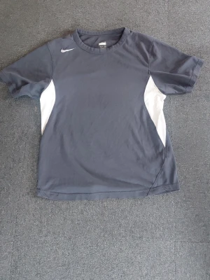 Mörkgrå Nike t-shirt med vita detaljer - Snygg mörkgrå t-shirt från Nike med vita paneler på sidorna och klassisk Nike-logga på bröstet. T-shirten har rund hals och korta ärmar, perfekt för en sportig look. Tillverkad i ett mjukt och lätt material som känns skönt mot huden.