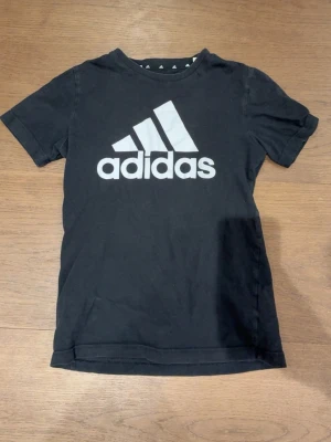 Svart Adidas t-shirt med logga - Snygg svart t-shirt från Adidas med stor vit logga på bröstet. Klassisk passform och rund halsringning. Perfekt till vardags för en sportig och avslappnad stil.