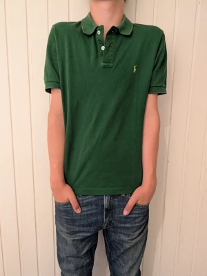 Grön pikétröja med krage från polo Ralph lauren - Snygg grön pikétröja med klassisk krage och korta ärmar. Tröjan har knappar vid halsen och en liten broderad logga på bröstet. Perfekt till jeans eller shorts för en avslappnad stil.