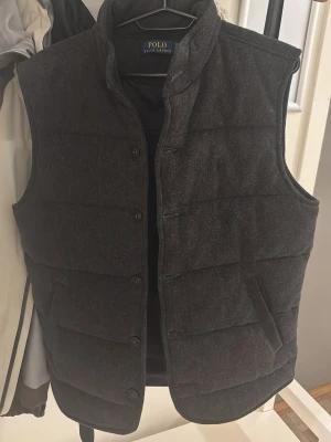 Mörkgrå dunväst från Polo Ralph Lauren - Snygg mörkgrå dunväst från Polo Ralph Lauren med ståkrage och diskreta sidofickor. Västen har en quiltad design och stängs med knappar framtill. Perfekt för lager på lager och ger en stilren look till din outfit. Den är helt ny 