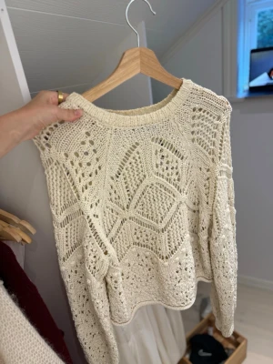 Vit virkad stickad tröja - Säljer en vit stickad tröja med virkat mönster och rund hals. Tröjan har långa ärmar och ett luftigt, genombrutet mönster som ger en trendig och avslappnad look. Perfekt att styla över ett linne eller t-shirt.