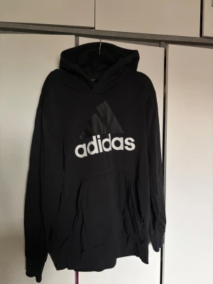 Svart hoodie från Adidas med logga - Snygg svart hoodie från Adidas med stor vit logga och klassiska tre ränder på bröstet. Lappen med storlek är borta men skulle uppskatta L/XL