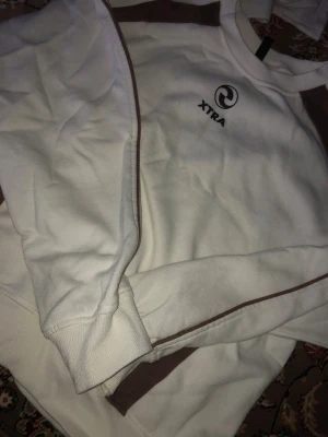 Vit och brun sweatshirt från XTRA - Säljer en vit sweatshirt från XTRA med bruna detaljer längs ärmar och sidor. Tröjan har rund halsringning, ribbade muddar och tryckt logga på bröstet. Perfekt för en avslappnad och sportig stil.