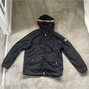 Svart vindjacka med huva - Snygg svart vindjacka med huva och dragkedja från moncler. Jackan har två framfickor med knappar och en detalj med röd, vit och blå rand på huvan. Liten patch på ena ärmen. Perfekt för blåsiga dagar och enkel att matcha med olika outfits. En av dragskedjorna är av men är en enkel fix