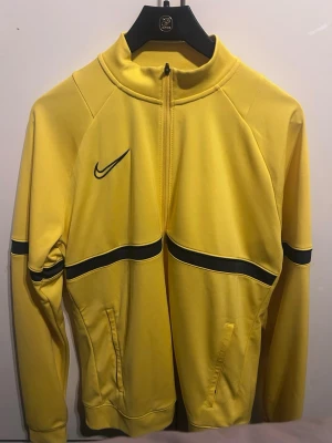 Gul Nike ziptröja med svarta detaljer - Gul ziptröja från Nike med svarta detaljer och logga på bröstet. Tröjan har hel dragkedja, ståkrage och två fickor framtill. Tillverkad i ett mjukt och sportigt material, perfekt för träning eller chill. Långärmad modell med svart rand över bröst och ärmar.