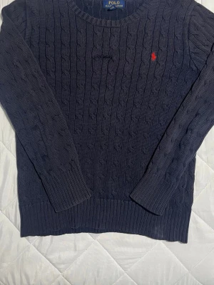 Polo Ralph Lauren Knitwear  - Säljer en klassisk och stilren Polo Ralph Lauren Knit i mörkblå färg med den ikoniska röda loggan. En tidlös tröja som passar perfekt till både vardag och mer uppklädda tillfällen.  Detaljer • Äkta Polo Ralph Lauren • Klassisk Cable Knit-design • Mörkblå med röd broderad logga • Mjuk och bekväm passform   Storlek: L youth som motsvarar S eller XS i vuxen storlek.  Skick: Använt skick. Tröjan har ett mindre hål framtill. Det finns också en liten fläck som går säkert att få bort.