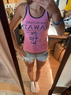 Rosa linne med Hawaii-tryck - Säljer ett rosa linne med mörkblå kant och tryck framtill med texten 'The Ultimate Hawaii Island Playground' samt palmer. Linnet är ärmlöst och har en avslappnad passform, perfekt för sommardagar.