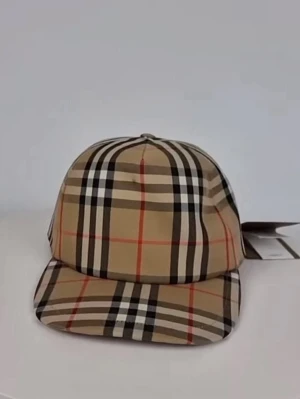 Beige burberry keps - Snygg keps i klassiskt beige rutmönster med svarta, vita och röda linjer. Kepsen har böjd skärm och justerbar baksida. Tillverkad i ett slätt tyg som ger en stilren look. Perfekt accessoar för att lyfta din outfit.