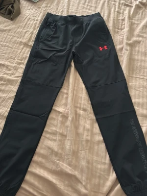 Svarta träningsbyxor Under Armour - Säljer ett par svarta träningsbyxor från Under Armour med röd logga på höften och diskret text längs benet. Byxorna har resår i midjan och vid bensluten samt två sidofickor. Perfekta för gymmet eller löpning.