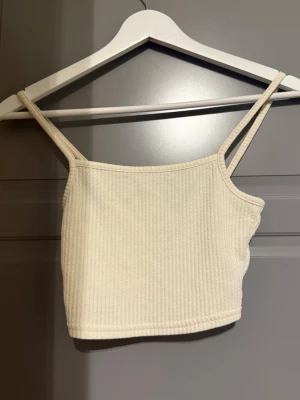 Vit ribbad croppad linnetopp - Säljer en vit croppad linnetopp med smala axelband och ribbad struktur. Toppen är ärmlös och har en enkel, stilren design som passar perfekt till jeans eller kjol. 